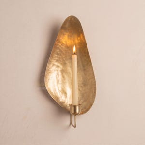 May include: A wall-mounted candle holder featuring a large, teardrop-shaped brass backplate with a hammered, textured finish. A single, slender, off-white taper candle sits upright in a small, cylindrical brass cup attached to the lower center.