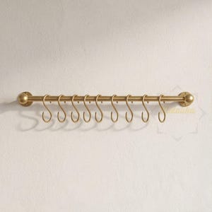 Op de afbeelding: Een horizontale metalen rail in messingkleur met een gladde cilindrische stang en afgeronde eindkappen. Negen bijpassende S-vormige haken hangen aan de stang, gelijkmatig verdeeld over de lengte. Het beslag is bedoeld voor wandmontage.