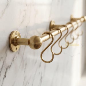 May include: A brushed brass wall-mounted rail featuring a cylindrical rod with rounded end caps and circular mounting plates secured by screws. Several matching S-shaped hooks are suspended from the horizontal bar for hanging various items.