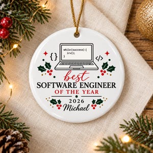 Peut inclure: Une décoration ronde en céramique blanche avec un graphique d'ordinateur portable noir affichant le code while(success) { irv(); } et le texte best SOFTWARE ENGINEER OF THE YEAR 2026 Michael, agrémenté de houx rouge, de feuilles vertes et d'or.