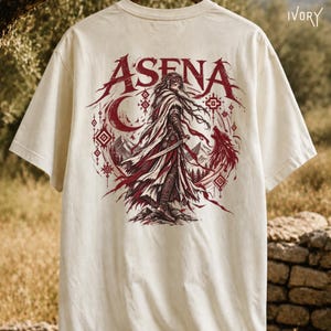 May include: An ivory t-shirt featuring a dark red graphic on the back. The design displays the text ASENA above a figure in flowing robes holding a sword, accompanied by a crescent moon, geometric patterns, mountains, and a wolf silhouette.