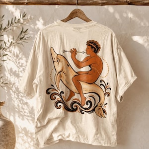 May include: An off-white short-sleeve t-shirt featuring a graphic print of a person playing a flute while riding a dolphin. The illustration uses shades of brown, black, and cream, incorporating swirling wave patterns and dotted lines.