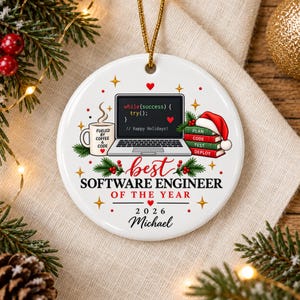 Può includere: Un ornamento rotondo in ceramica bianca con un computer portatile che mostra codice, una tazza di caffè con l'etichetta FUELED BY COFFEE AND CODE e una pila di libri etichettati PLAN, CODE, TEST, DEPLOY. Il testo recita BEST SOFTWARE ENGINEER OF THE YEAR 2026 Michael.