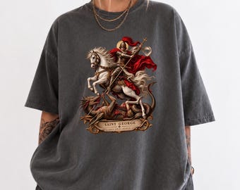 May include: A charcoal gray short-sleeve t-shirt featuring a graphic print of Saint George on a white horse battling a dragon. The design includes a red cape, gold armor, and a banner at the bottom with the text SAINT GEORGE ORA PRO NOBIS.