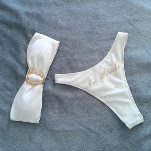 May include: A white bikini bottom featuring the text Mrs. Borselli in gold script and a matching white fabric band cinched with a textured gold-toned metal ring. The set is made of a soft, white fabric with a subtle ribbed texture throughout.