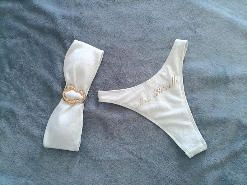 May include: A white bikini bottom featuring the text Mrs. Borselli in gold script and a matching white fabric band cinched with a textured gold-toned metal ring. The set is made of a soft, white fabric with a subtle ribbed texture throughout.