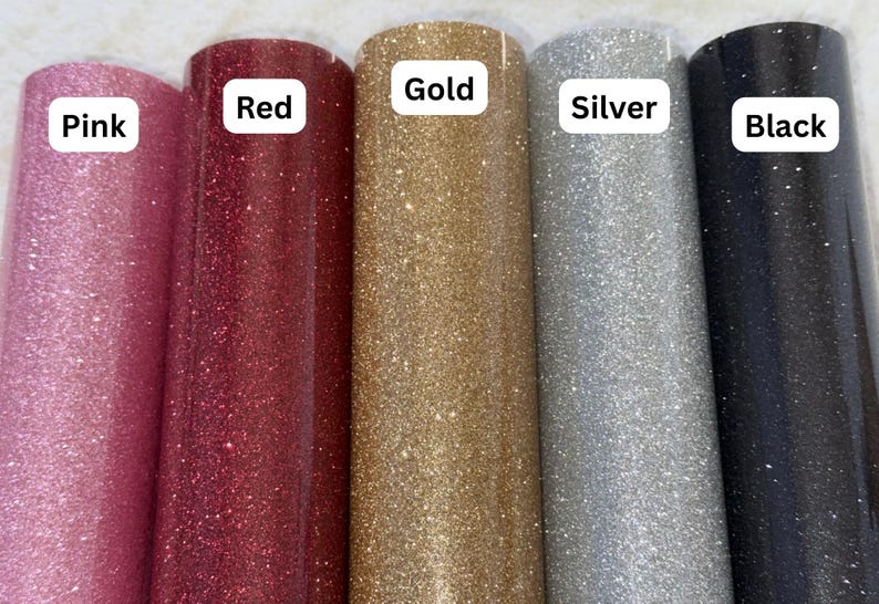 May include: Five rolls of glitter adhesive vinyl arranged side by side in pink, red, gold, silver, and black. Each roll features a white rectangular label with the corresponding color name printed in black text: Pink, Red, Gold, Silver, and Black.