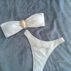 May include: A white two-piece bikini set featuring a strapless bandeau top with a central gold-toned irregular ring accent and high-cut bikini bottoms embroidered with the gold text Mrs. Borselli on the front hip area.