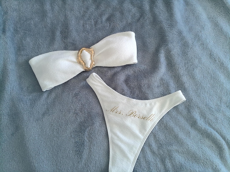 May include: A white two-piece bikini set featuring a strapless bandeau top with a central gold-toned irregular ring accent and high-cut bikini bottoms embroidered with the gold text Mrs. Borselli on the front hip area.