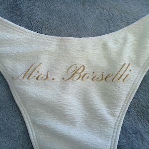 May include: A white textured bikini bottom featuring the text Mrs. Borselli printed in a gold glitter script font across the center. The garment has a high-cut leg design and a soft, crinkled fabric texture throughout the entire piece.
