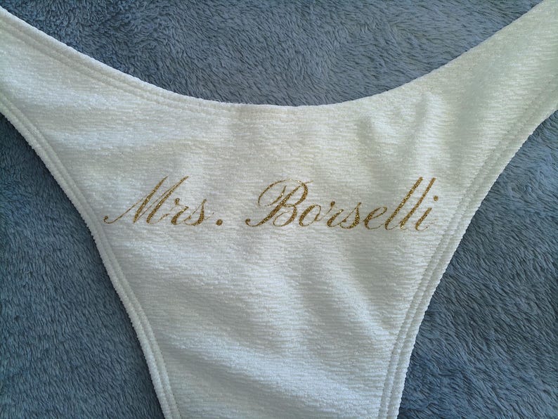 May include: A white textured bikini bottom featuring the text Mrs. Borselli printed in a gold glitter script font across the center. The garment has a high-cut leg design and a soft, crinkled fabric texture throughout the entire piece.