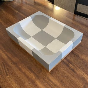Op de afbeelding: Een rechthoekig dienblad met een dambordpatroon van afwisselend lichtgrijze en witte vierkanten. Het dienblad heeft een hol, komvormig midden met afgeronde randen en een platte basis, afgewerkt met een glad oppervlak en een lichtgekleurde houten onderrand.