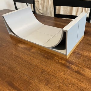Peut inclure: Une rampe de fingerboard en demi-lune gris clair avec une surface incurvée lisse et des parois latérales verticales. La structure est montée sur une base rectangulaire beige solide, offrant une plateforme stable pour les figures de skate.