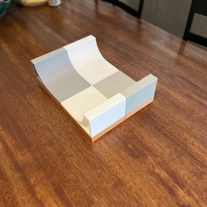 Peut inclure: Un présentoir rectangulaire doté d'une surface supérieure incurvée et concave avec un motif en damier composé de carrés alternés gris clair et blancs. La base du présentoir est fabriquée dans un matériau en bois clair, offrant une assise stable.