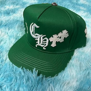 May include: Green baseball cap featuring white embroidered lettering and cross motifs. The cap has a flat peak and a small silver crown detail. The text on the cap is in a decorative style.