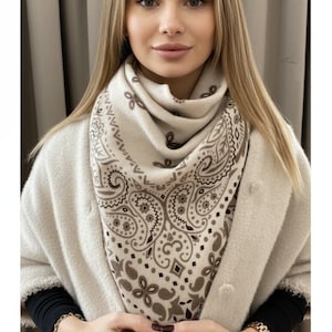 Peut inclure: Un foulard carré présentant une base de couleur crème avec un motif cachemire et géométrique marron. Le foulard est plié en forme de triangle autour du cou, mettant en valeur un imprimé détaillé avec des volutes, des points et des losanges.