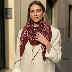 Op de afbeelding: Een vierkante sjaal in donkerbordeaux met een lichtroze paisley- en bloemenpatroon, voorzien van een gerafelde rand. De sjaal wordt gedragen over een lichtbeige blazer met één knoop, lange mouwen met omgeslagen boorden en twee voorzakken.