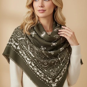 Peut inclure: Un foulard carré vert olive orné d'un motif cachemire et floral blanc avec une bordure décorative. Le foulard est drapé sur les épaules et présente une texture tissée douce avec un bord fini, mettant en valeur des motifs symétriques.