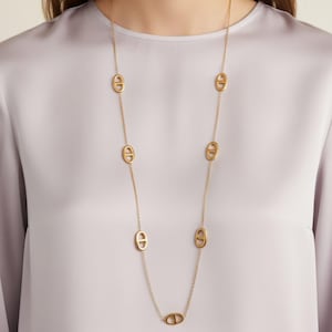Peut inclure: Un long collier doré avec des maillons ovales espacés le long de la chaîne. Le collier est porté sur un chemisier violet clair. Les maillons ont un design distinctif, ajoutant une touche d'élégance au bijou.