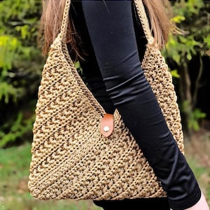 Peut inclure: Un sac à bandoulière beige crocheté avec une fermeture à lanière en cuir marron. Le sac a une texture tissée et une bandoulière tressée. Le sac est porté sur un haut noir à manches longues.