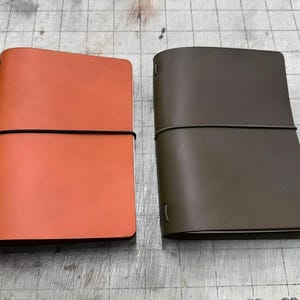 以下が含まれることがあります： オレンジ色とオリーブグリーンの2冊の革製ジャーナルが並んで表示されています。各ジャーナルは黒いゴムバンドで閉じられています。ジャーナルは、グリッドパターンのある灰色の表面にあります。