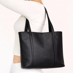May include: A black tote bag with two vertical seams on the front. The bag has two long handles and is carried over the shoulder. The bag is made of a smooth material and has a rectangular shape.
