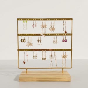 May include: A gold-colored metal earring display stand with three horizontal bars, holding various earrings. The stand is mounted on a light-colored wooden base. The earrings include pearl, crystal, and floral designs.