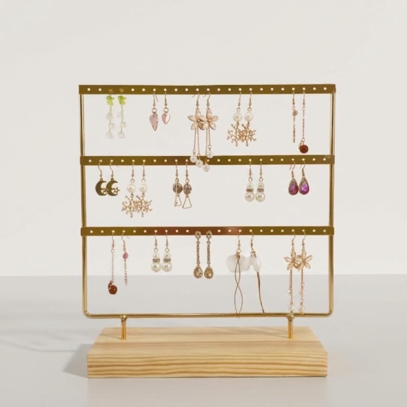 May include: A gold-colored metal earring display stand with three horizontal bars, holding various earrings. The stand is mounted on a light-colored wooden base. The earrings include pearl, crystal, and floral designs.