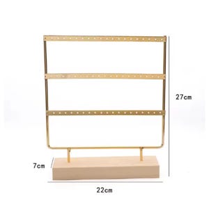 May include: A gold-colored metal earring display stand with three horizontal bars, each with multiple holes for earrings. The stand is 27 cm tall, 22 cm wide, and has a 7 cm wooden base.