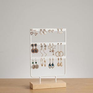 May include: A white metal earring display stand with multiple pairs of earrings. The stand has a rectangular wooden base and a grid-like structure to hold the earrings. Various earring styles include studs, hoops, and dangling designs in gold, silver, and blue.