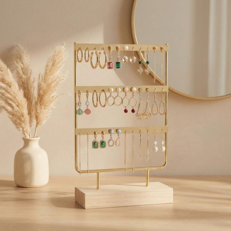 May include: A gold-colored earring display stand with three horizontal bars, holding various earrings. The stand is on a light wood base. A vase with dried plants is to the left, and a round mirror is in the background.