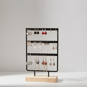 May include: Black metal earring display stand with three horizontal bars, holding various earrings. The stand is mounted on a light wood base. Earrings include studs, hoops, and dangling styles in silver, gold, and pearl.