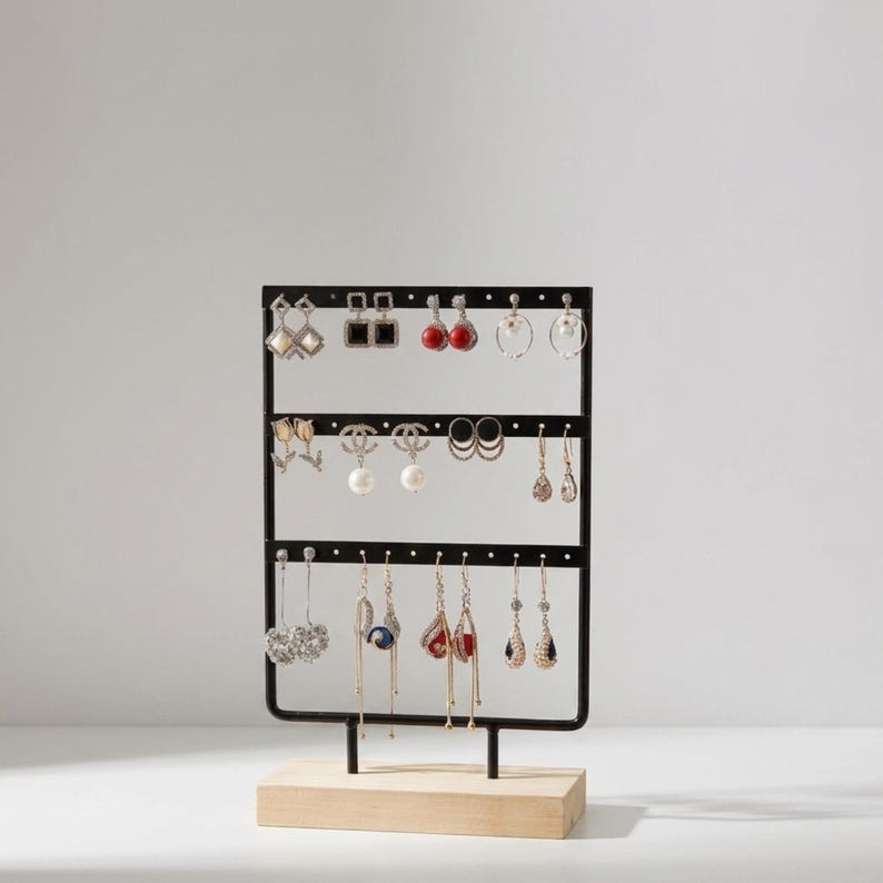 May include: Black metal earring display stand with three horizontal bars, holding various earrings. The stand is mounted on a light wood base. Earrings include studs, hoops, and dangling styles in silver, gold, and pearl.