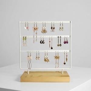 May include: A white metal earring display stand with three horizontal bars, holding various earrings. The stand sits on a light wood base. The earrings include pearl, floral, and dangling designs, showcasing a variety of colors and styles.