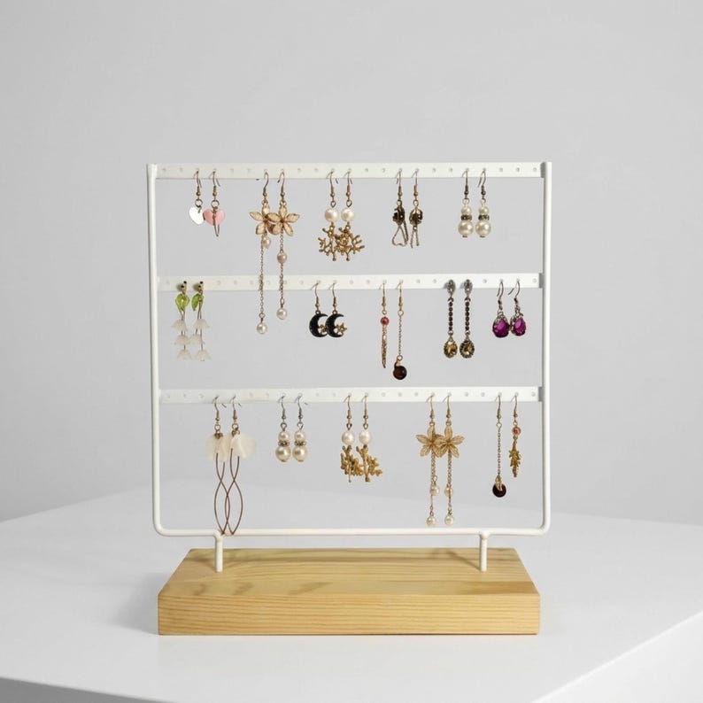 May include: A white metal earring display stand with three horizontal bars, holding various earrings. The stand sits on a light wood base. The earrings include pearl, floral, and dangling designs, showcasing a variety of colors and styles.