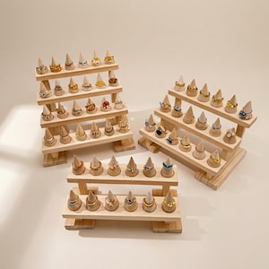 May include: Three tiered wooden ring display stands with multiple conical pegs. The stands are arranged in a group, holding various metal rings in gold, silver, and rose gold tones, some featuring small colorful stones or decorative shapes.