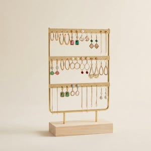 May include: A gold-colored metal earring display stand with three tiers, holding various earrings with colorful gemstones and pearls. The stand is mounted on a light-colored wooden base, showcasing a collection of jewelry.