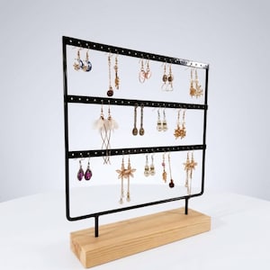 May include: A black metal earring display stand with three horizontal bars and a wooden base. The stand holds various earrings of different styles and colors, including gold, silver, and purple. The earrings are displayed against a white background.