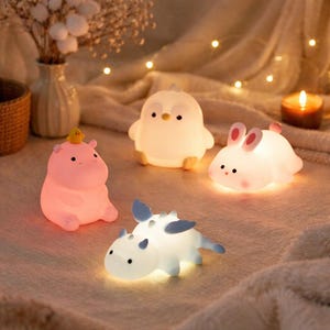 Peut inclure: Ensemble de veilleuses lumineuses en forme de personnages de dessins animés. L'ensemble comprend un hippopotame rose avec un canard jaune, un pingouin blanc, un lapin blanc aux oreilles roses et un dinosaure bleu et blanc. Les lumières émettent une lueur douce et chaleureuse.