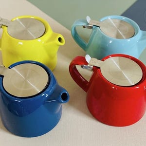Peut inclure: Quatre petites théières colorées en jaune, bleu, rouge et turquoise. Chaque théière est équipée d'un infuseur en acier inoxydable et d'une poignée incurvée. Les théières sont disposées sur une surface neutre, avec un design minimaliste.