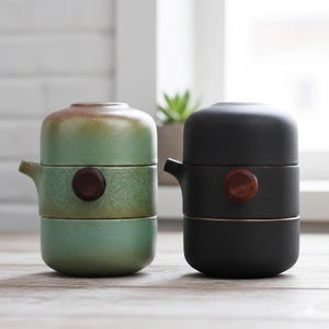 Peut inclure: Deux services à thé en céramique. L'un est vert avec un bouton et un bec verseur marron, l'autre est noir avec un bouton et un bec verseur en bois. Les deux ensembles sont empilés, avec un couvercle, une section centrale et une base.