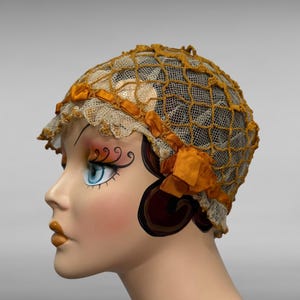 May include: A vintage cloche hat. The hat has a net-like design in a golden-yellow colour, with a cream-coloured lace trim and small orange-yellow bows. The hat is displayed on a mannequin head with painted features.