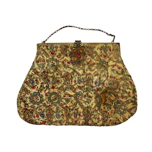 May include: A gold-toned fabric handbag featuring an intricate floral and vine pattern in shades of red, blue, and green. The bag has a metal frame, a thin chain handle, and a central rectangular clasp adorned with a dark amber-colored stone.