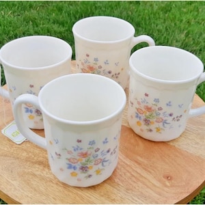 May include: Four white ceramic mugs with floral patterns in pink, blue, and yellow. Each mug has a curved handle and is placed on a round wooden surface. The background is green grass.