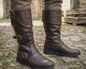 Könnte beinhalten: Braune kniehohe Lederstiefel mit dekorativen Schnallen. Die Stiefel haben einen umgeklappten oberen Teil und eine flache Sohle. Die Schnallen sind bronzefarben. Die Stiefel werden mit einer braunen Hose getragen.