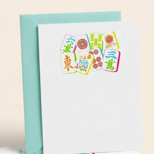 May include: A white greeting card with a teal envelope. The card features colorful illustrations of Mahjong tiles with Chinese characters and floral designs. The tiles are in shades of green, pink, yellow, and orange.