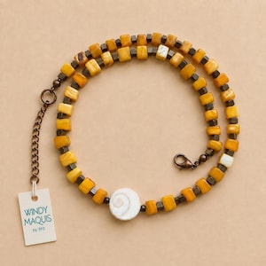 May include: A necklace featuring alternating amber-colored square beads and small metallic cubes, centered with a white spiral shell disc. The piece includes a bronze-toned chain extender and a lobster clasp, with a tag reading WINDY MAQUIS by 393.