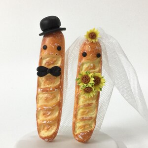 Puede incluir: Un adorno para tarta de boda caprichoso con dos figuras de baguette. Una baguette lleva un sombrero de copa negro y una pajarita, la otra un velo y girasoles. Las figuras están sobre una base circular blanca.