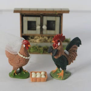 Puede incluir: Un conjunto de figuras caprichosas con temática de granja que presenta una gallina con velo y un gallo con pajarita, de pie frente a un gallinero en miniatura. También hay una pequeña cesta de huevos.