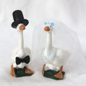 May include: Two white goose figurines standing on green bases. One goose wears a black top hat and a black bow tie. The other goose wears a white tulle veil with a small blue beaded crown and a matching light blue beaded necklace around its neck.